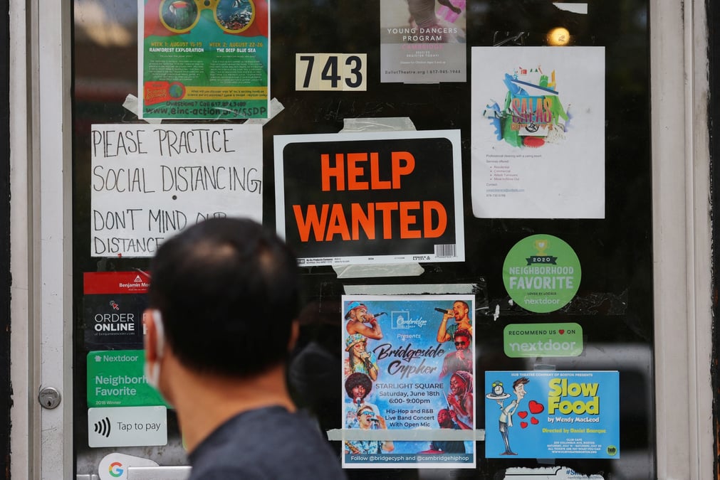 Photo d'un pieton qui passe devant un panneau "help wanted" a la porte d'une quincaillerie