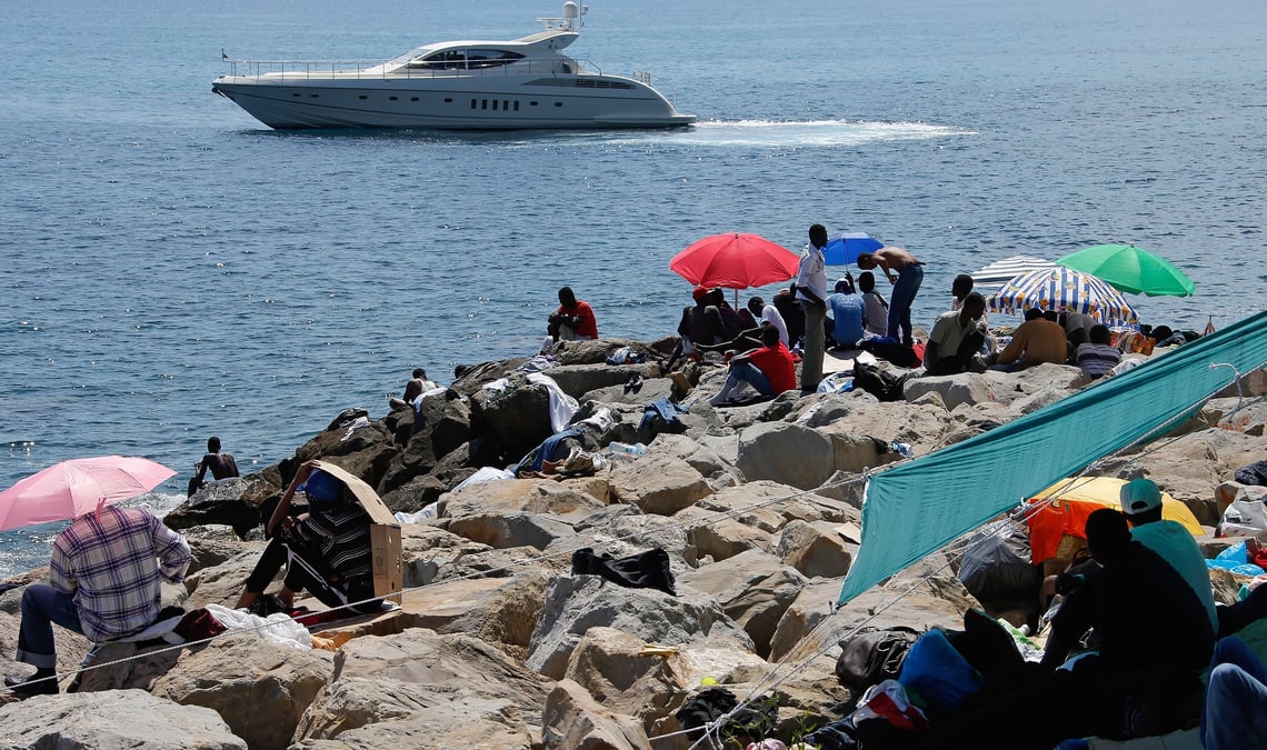 Un bateau de luxe passe devant un groupe de migrants installes sur les rochers de la digue a la frontiere de saint ludovic, en mediterranee, entre vintimille et menton