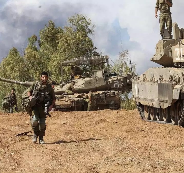 Le char Merkava IV apparaissant à l'arrière-plan est surmonté d'une sorte de cage protégeant la partie supérieure du véhicule.