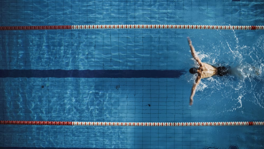 Aerial Top View Male Swimmer Swimming in Swimming Pool. Professional Determined Athlete Training for the Championship, using Butterfly Technique. Top View Shot - Aerial Top View Male Swimmer Swimming in Swimming Pool. Professi