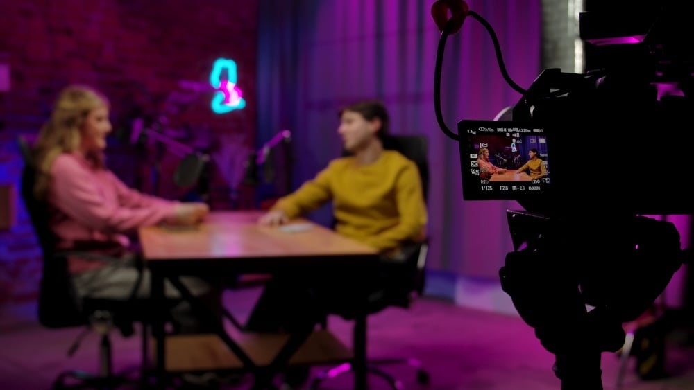 Online podcast. Backstage blurred shot of host and guest recording interview in loft podcast studio, creating digital content, camera on tripod filming at side, display pov. - Backstage blurred shot of host and guest recording interview in loft podcast...