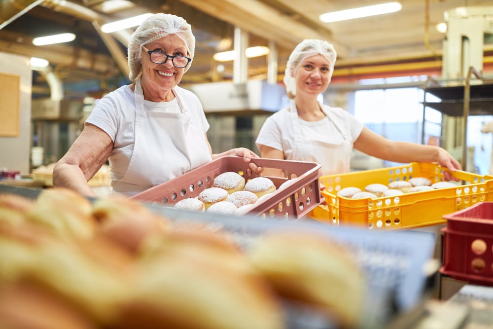 Two female bakers carry freshly baked Berlin pancakes in boxes in a large bakery - Bakers carry fresh Berlin pancakes in boxes