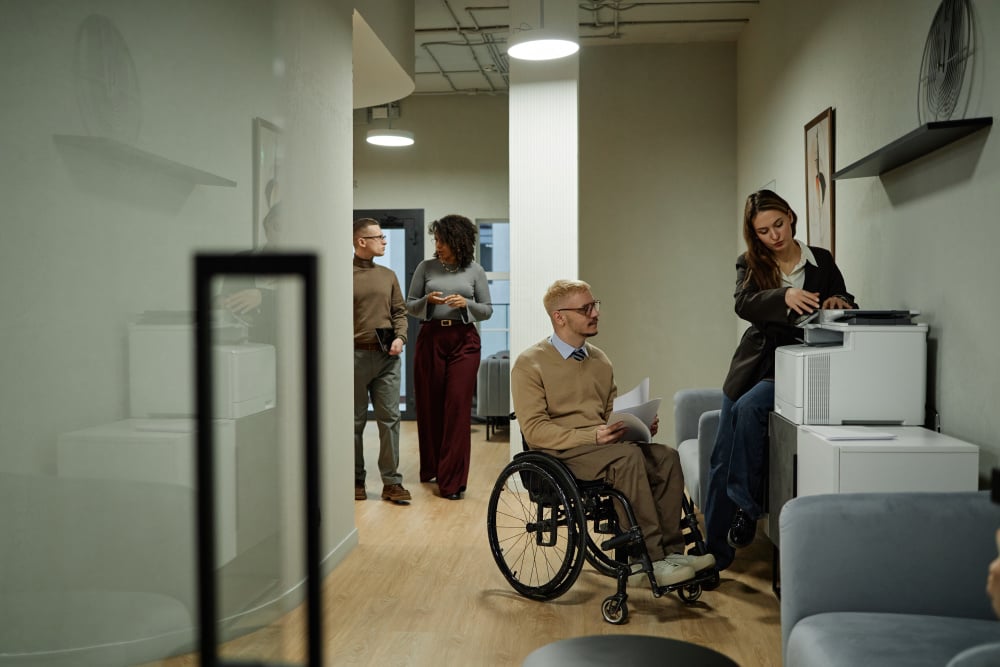 Caucasian young adult man with disability using wheelchair holding documents near printer while woman operating printer and multiethnic colleagues walking in office hallway - Caucasian Young Adult Man with Disability Using Wheelchair in Modern Office -...