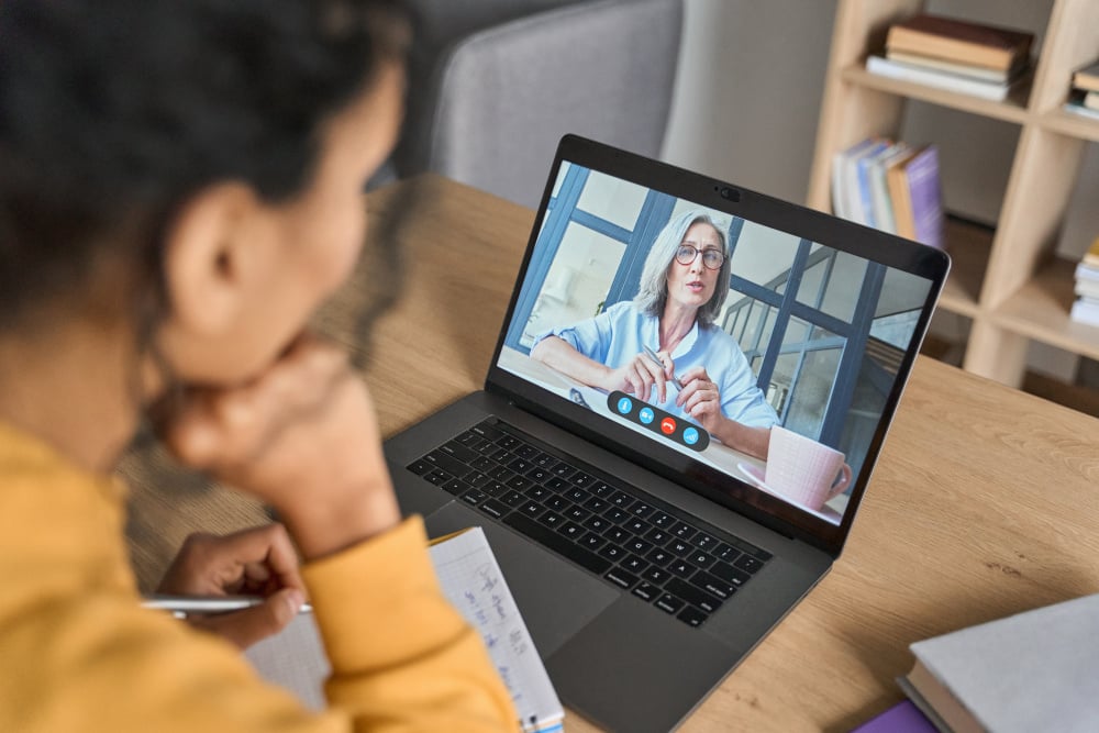 Over shoulder view of female adult student having virtual meeting online call educational webinar chatting with senior teacher at home office writing notes. Video e learning conference call on pc. - African American female student having video...