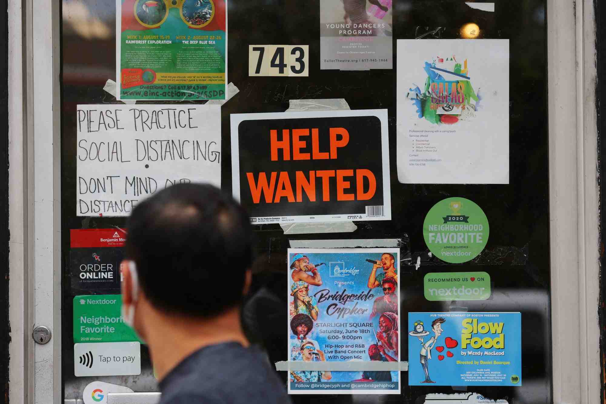 Photo d'un pieton qui passe devant un panneau "help wanted" a la porte d'une quincaillerie