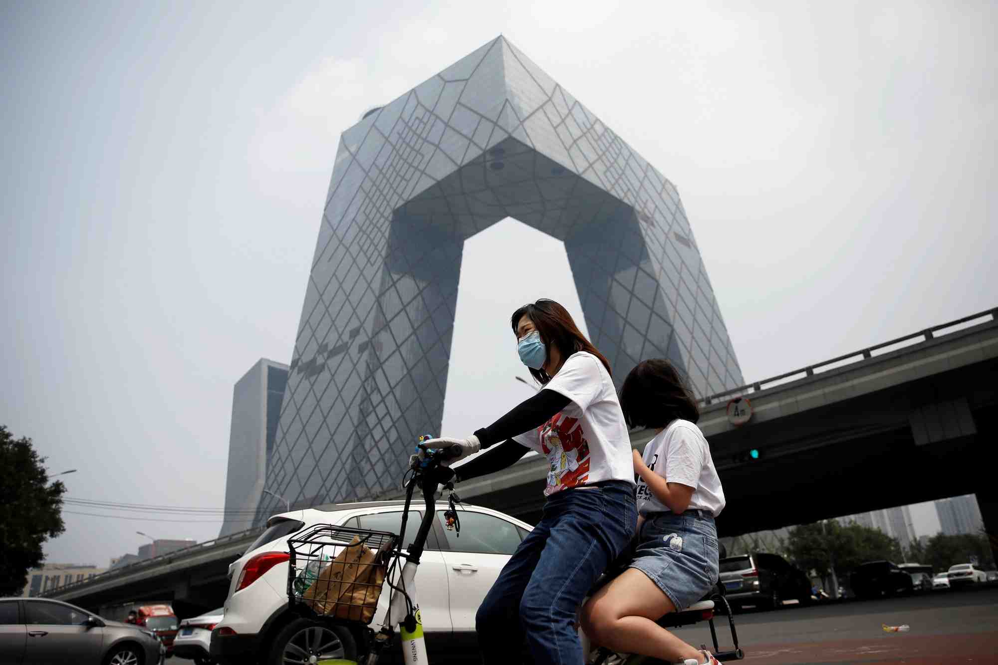 Chine : une femme portant un masque roule en vélo électrique devant le siège de la Télévision centrale de Chine à Pékin le 16 juillet 2020