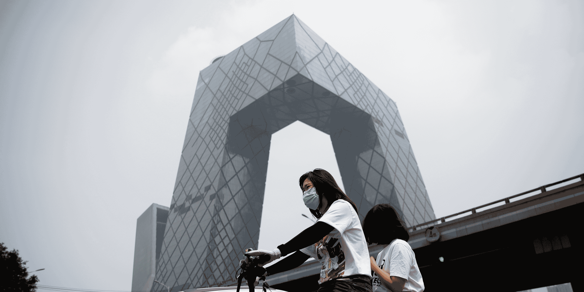 Chine : une femme portant un masque roule en vélo électrique devant le siège de la Télévision centrale de Chine à Pékin le 16 juillet 2020