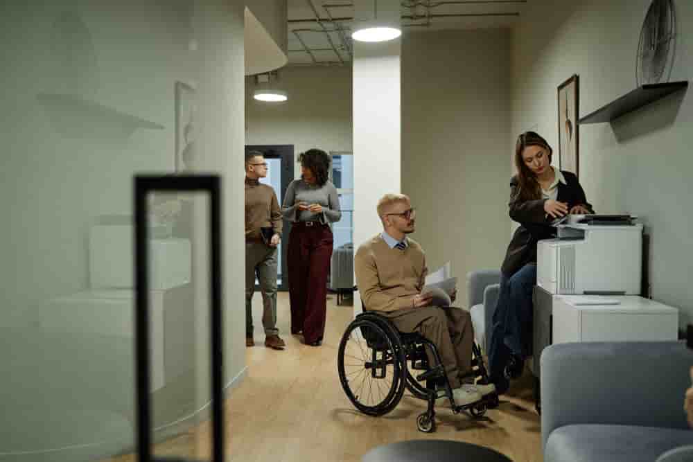 Caucasian young adult man with disability using wheelchair holding documents near printer while woman operating printer and multiethnic colleagues walking in office hallway - Caucasian Young Adult Man with Disability Using Wheelchair in Modern Office -...