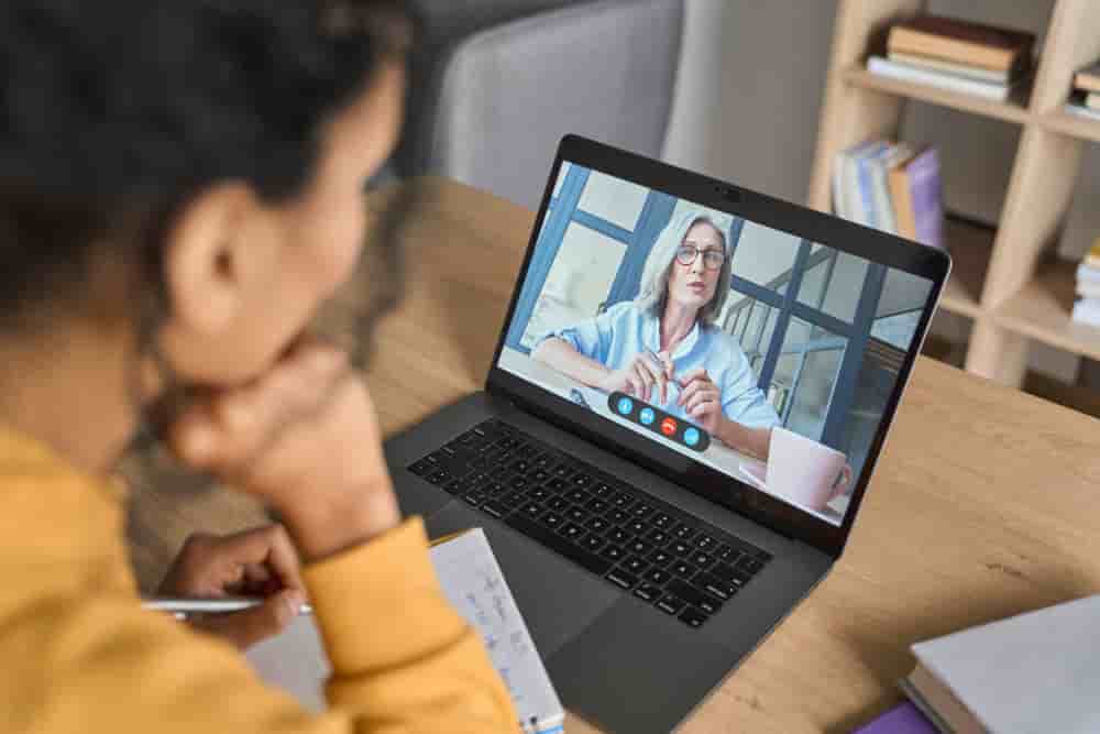 Over shoulder view of female adult student having virtual meeting online call educational webinar chatting with senior teacher at home office writing notes. Video e learning conference call on pc. - African American female student having video...