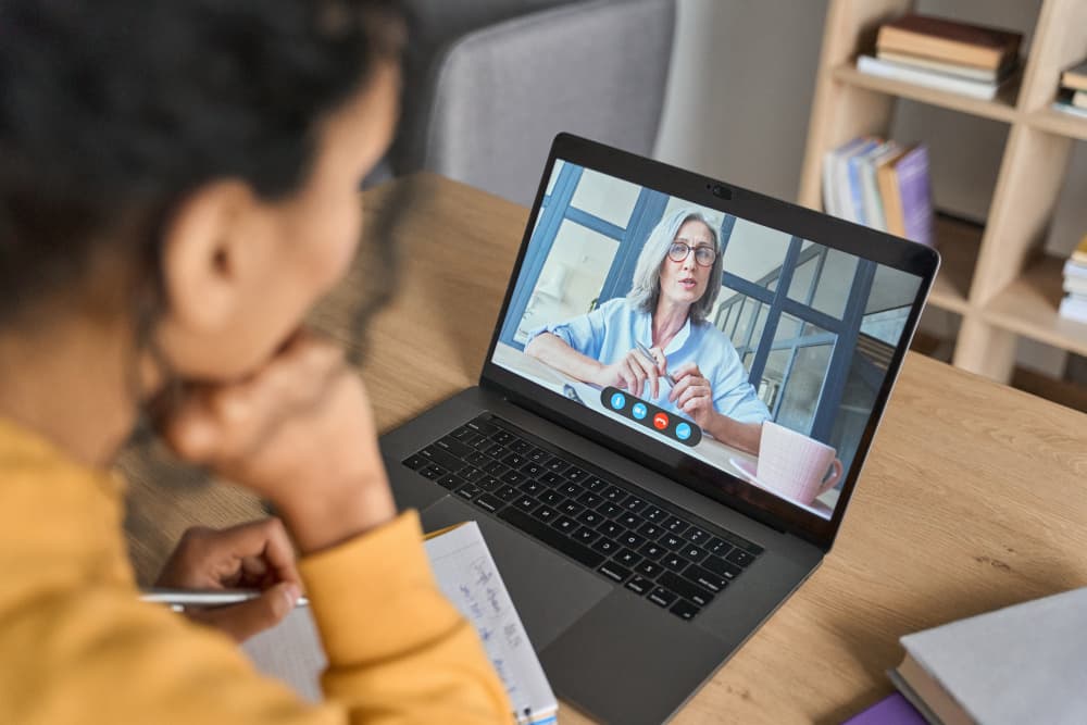 Over shoulder view of female adult student having virtual meeting online call educational webinar chatting with senior teacher at home office writing notes. Video e learning conference call on pc. - African American female student having video...